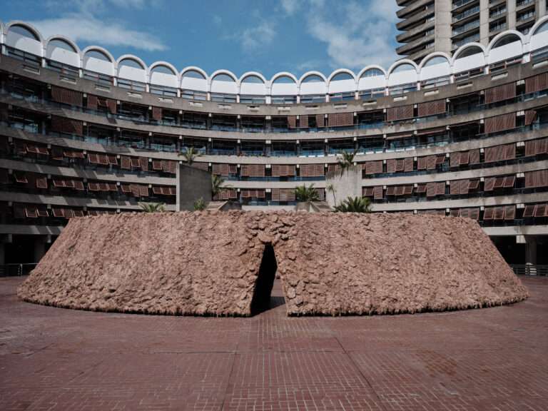 An earthen circular installation sits in front of a curved brutalist concrete building facade.
