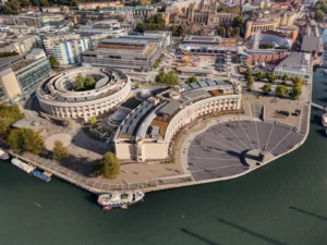Aerial photograph showing a large circular building massing and a curved crescent building facing a public waterfront plaza.