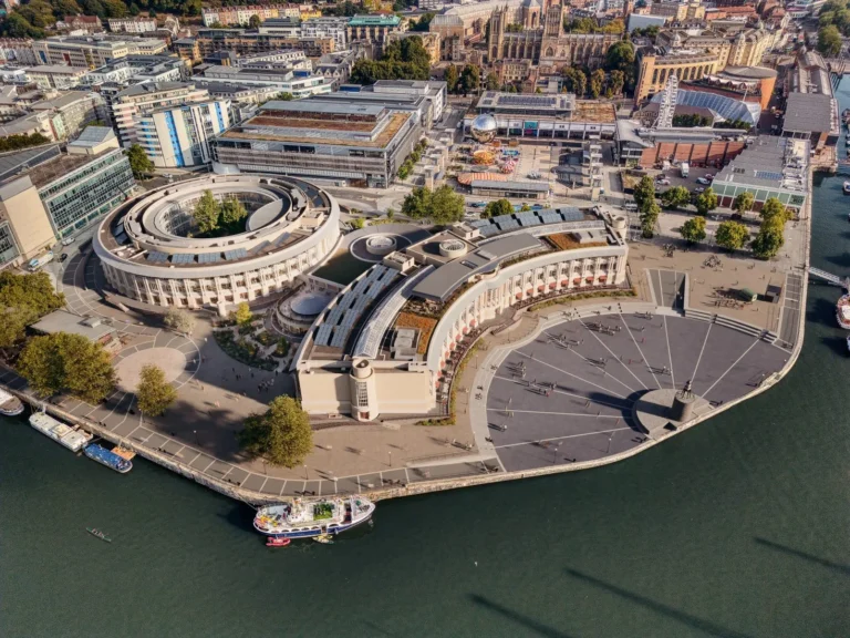 Aerial photograph showing a large circular building massing and a curved crescent building facing a public waterfront plaza.