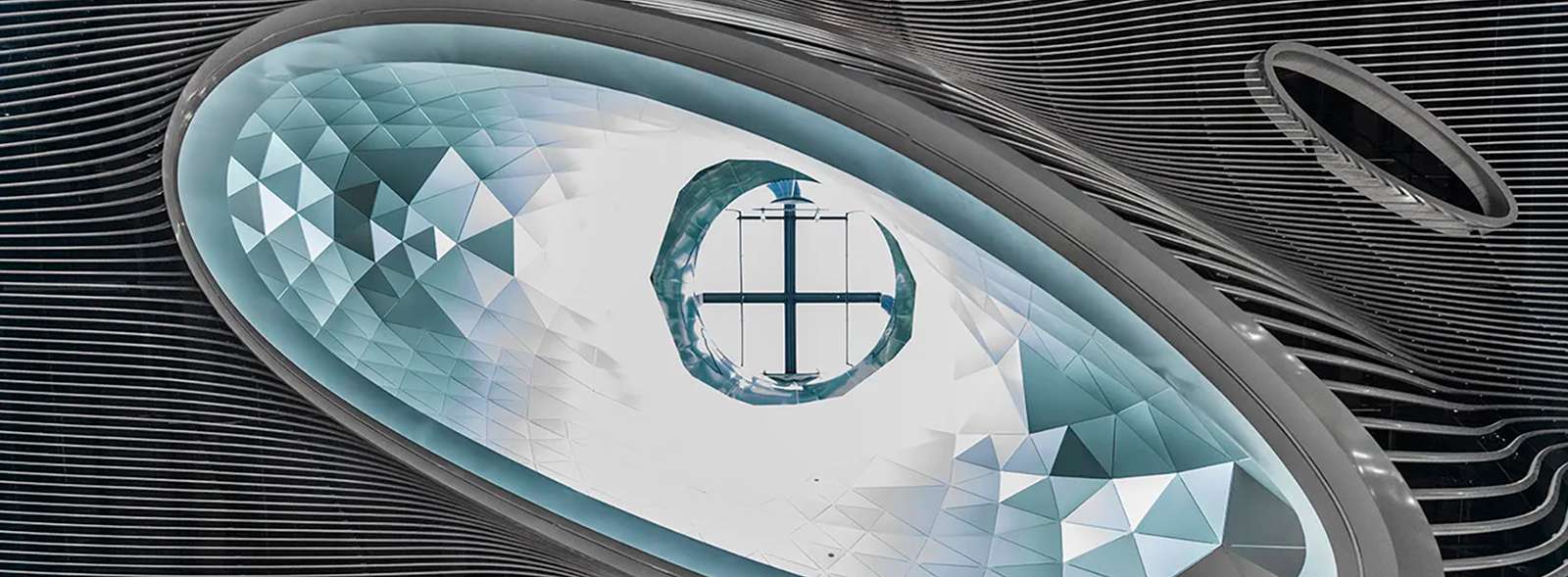 A close-up view of an undulating ceiling system with an oval skylight opening and geometric aluminum paneling.