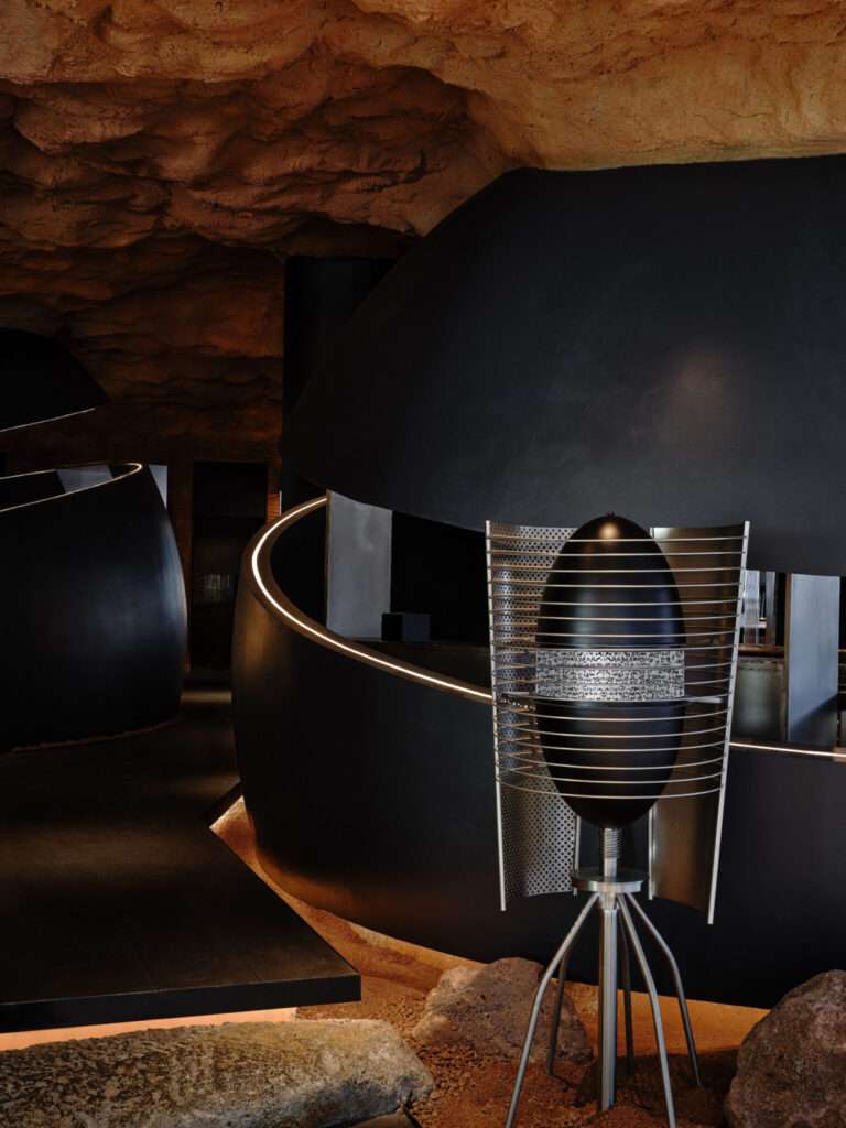 Close-up of an egg-shaped black and metallic decorative capsule on a tripod stand with illuminated typographic details in a cave-like interior path.