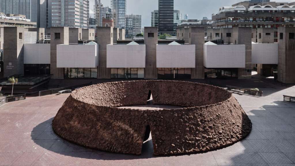 An elevated view of a wide earthen circular structure with an open central void in a concrete plaza.