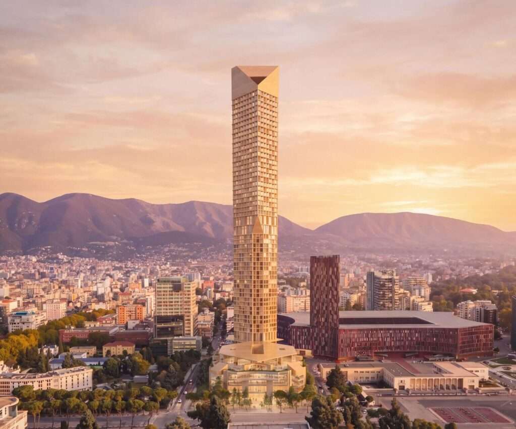 Aerial view of a tall, geometric tower rising above a city with mountains in the background.