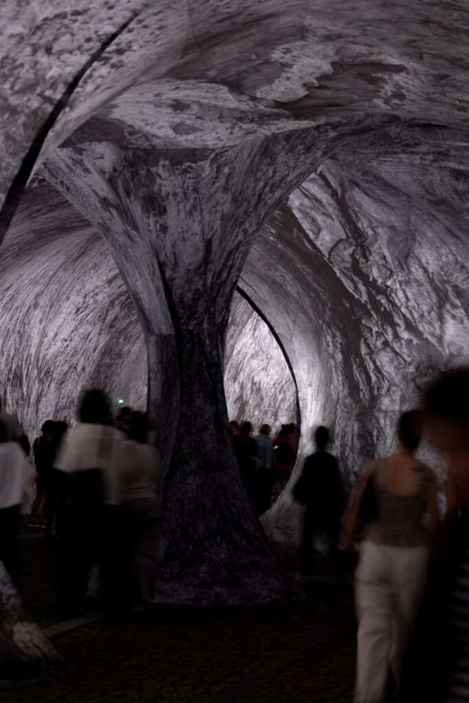 A blurred crowd of visitors walking around a large, central fabric column supporting a cave-like ceiling.