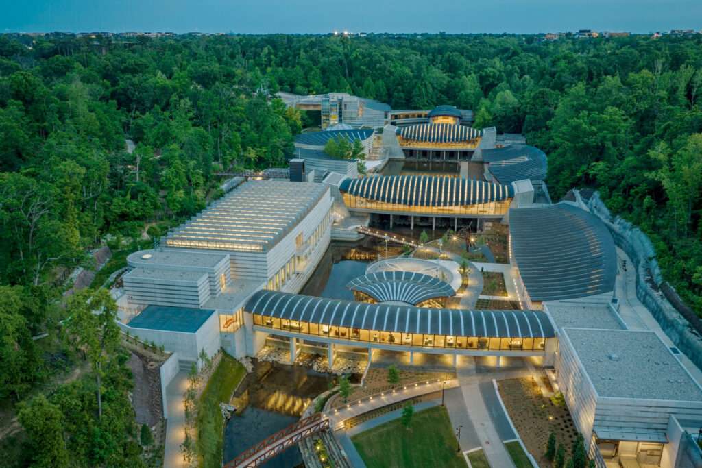 An aerial perspective of a U-shaped museum campus with curved and sawtooth roofs nestled in a dense forest.