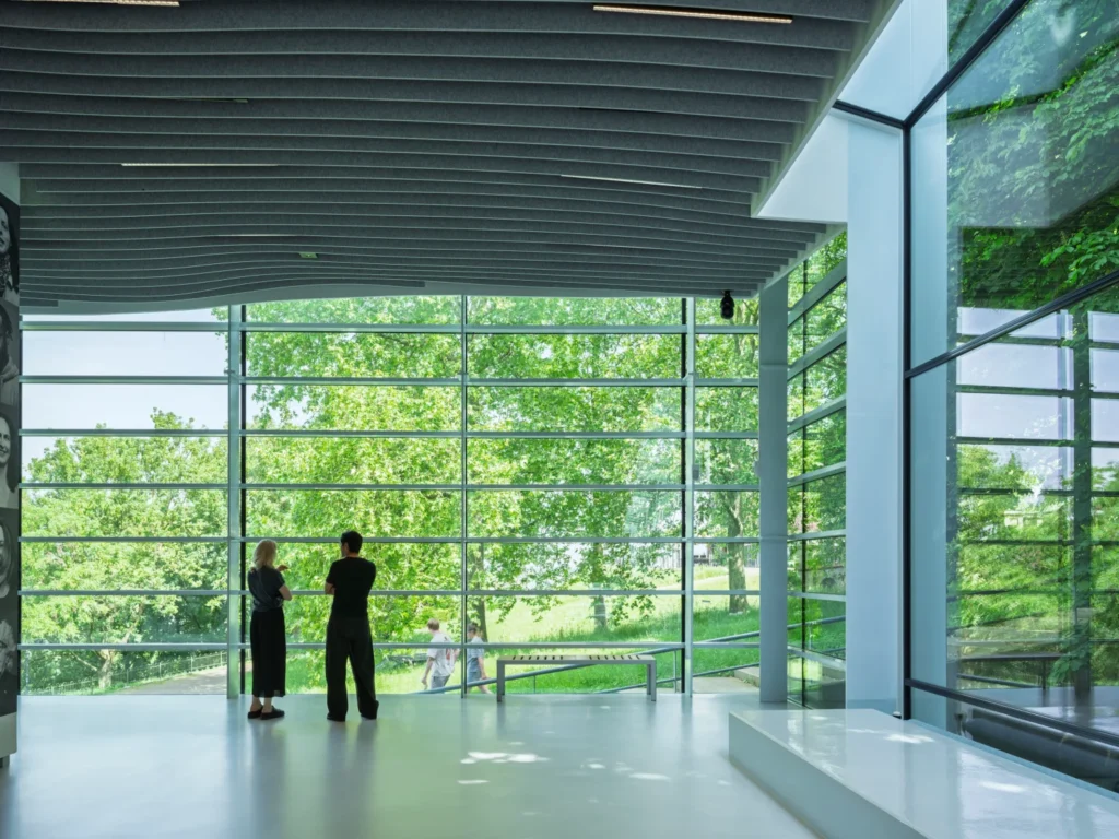 Interior perspective looking through a large glass facade towards a green landscape edge with trees.