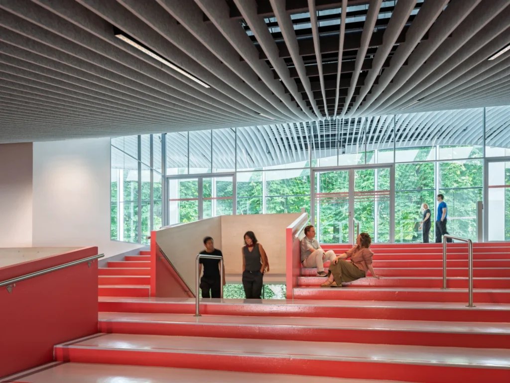 Interior view of a wide central staircase with bright red steps and a slatted acoustic ceiling.