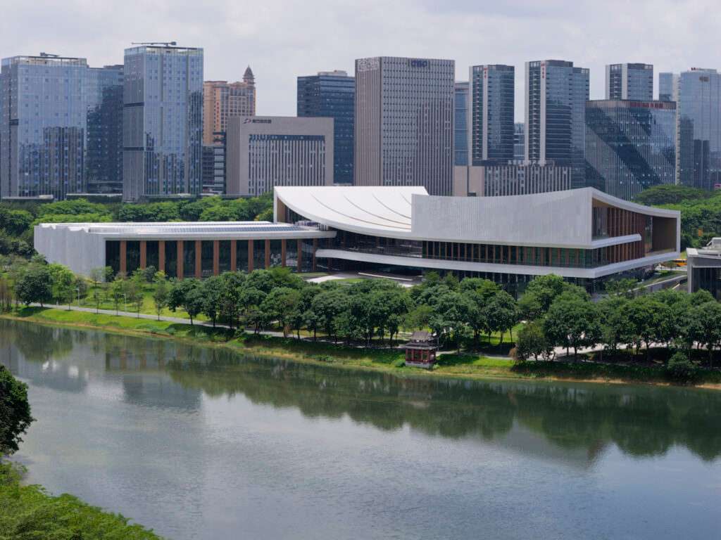 A wide view of the modern white building across a body of water with high-rise buildings in the background