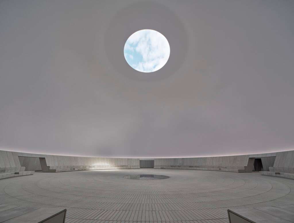 An interior shot of a massive concrete circular hall under a dome with a single round oculus framing a blue sky with clouds.