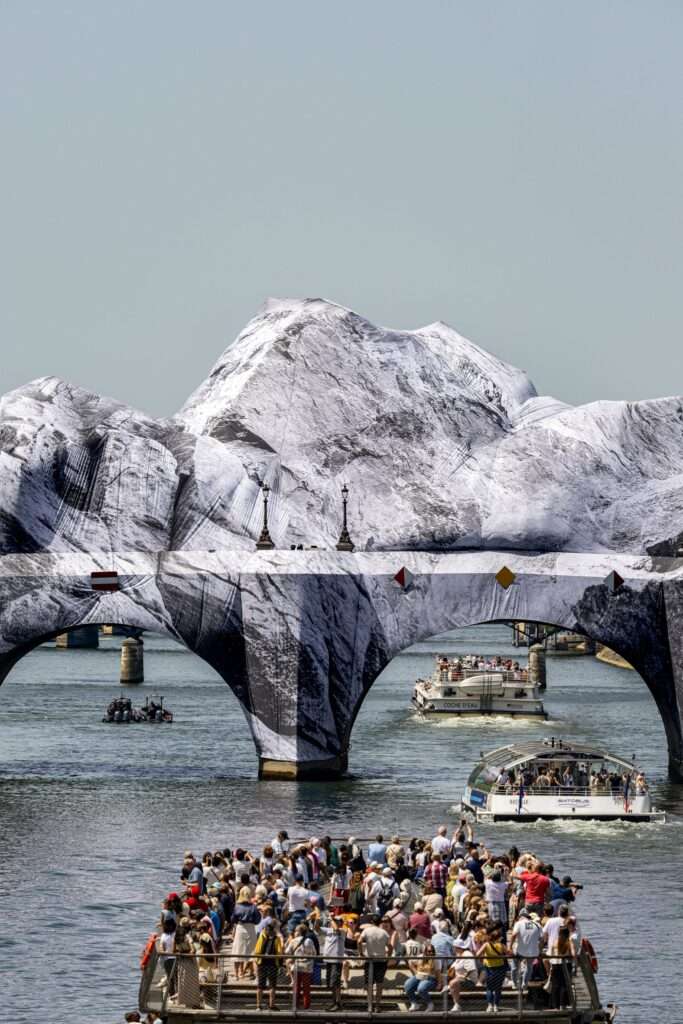 An arched river bridge covered with a temporary fabric installation shaped like mountain peaks, with tourist boats below.