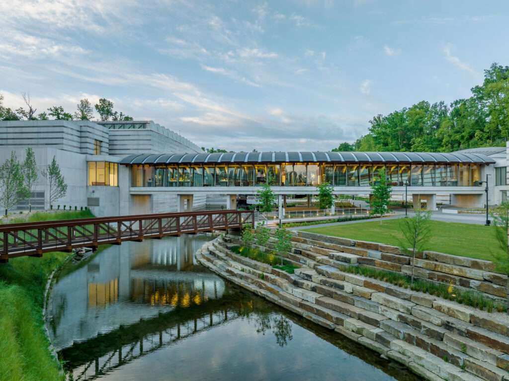 A long glass-enclosed bridge gallery stands elevated above a green lawn, pedestrian path, and stone waterfront.