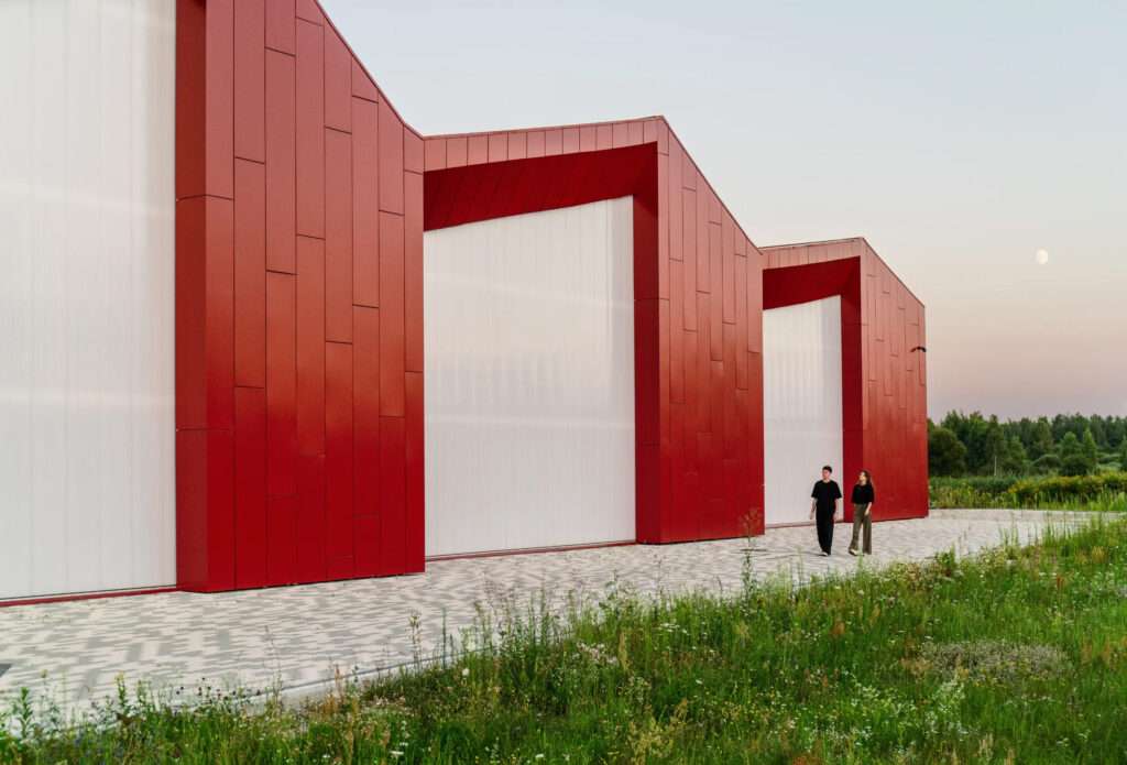 Two people walking on a paved path next to a red panel clad building with translucent sections.