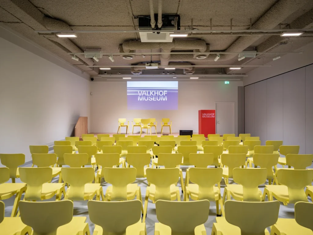 An auditorium interior with neat rows of yellow chairs facing a projection screen on a stage.