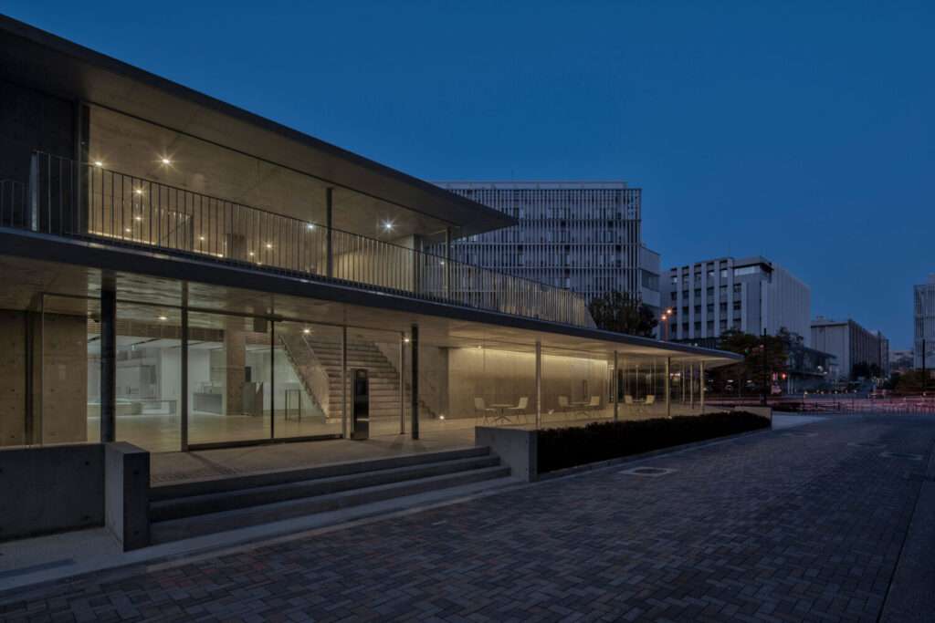 A dusk view of a concrete and glass building with illuminated interiors facing a paved stone path.