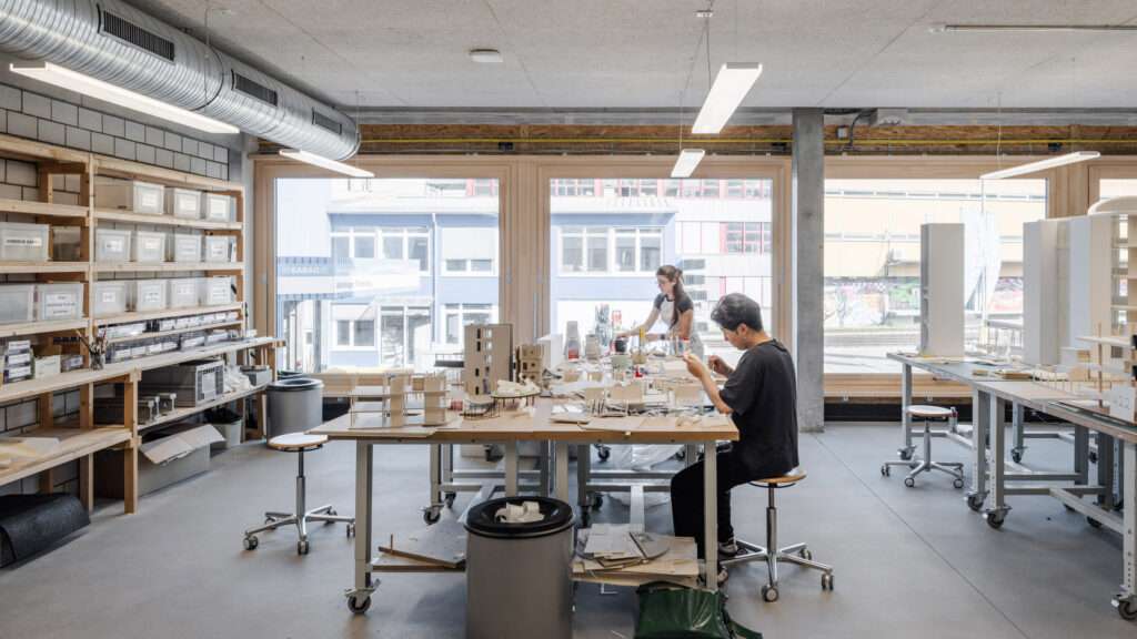 An architectural workshop space with long model-making tables, concrete walls, and large windows.