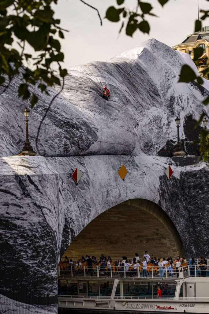 A technician wearing a safety helmet works on the side of a massive fabric mountain installation draped over a bridge.