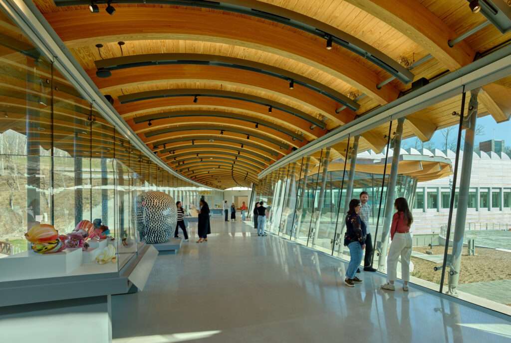 Interior view of a long hallway with a barrel-vaulted timber ceiling and floor-to-ceiling glass windows.