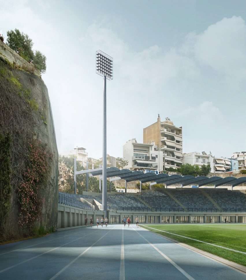 Runners on a blue athletic track next to a high rock wall and stadium seating.