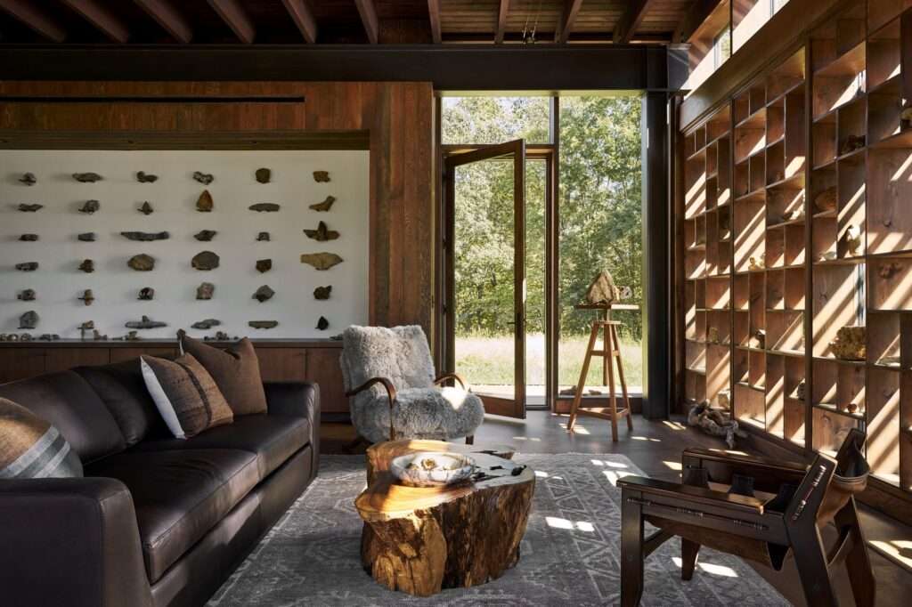 Interior view of Rock Pavilion featuring a leather sofa, a raw wood log coffee table, an open wooden shelving unit holding geological stones, and sunlight casting linear shadows.