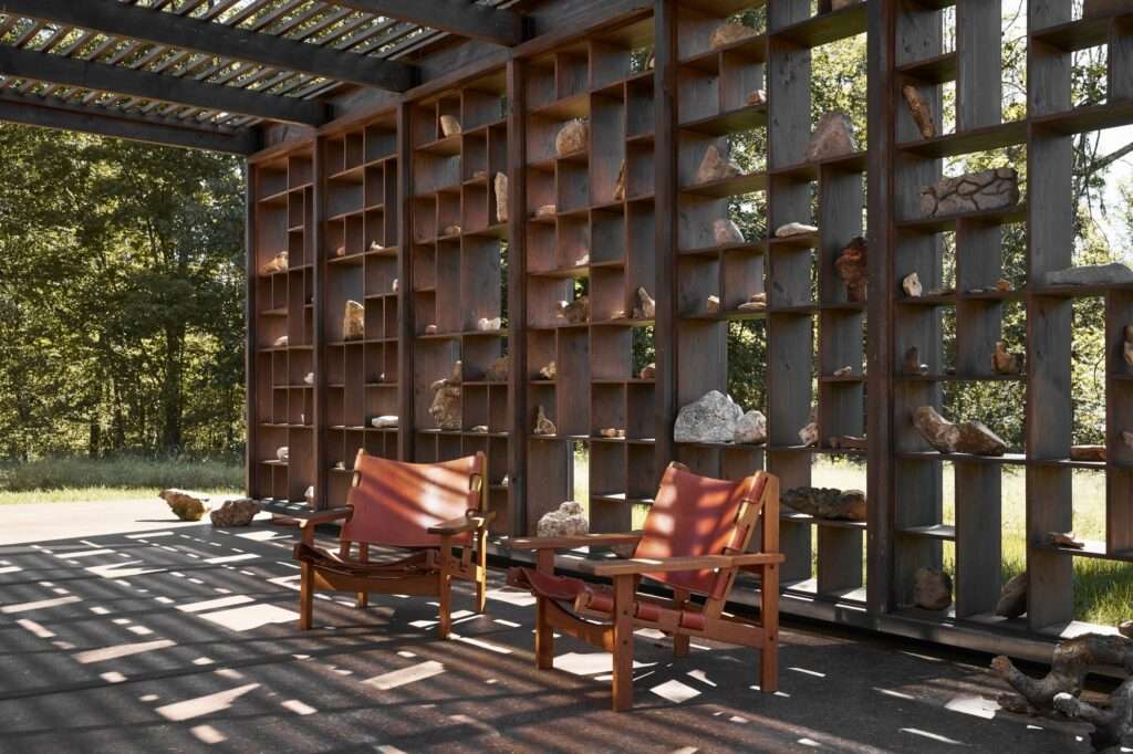 Outdoor dining terrace of the Rock Pavilion with two leather lounge chairs under a dark timber trellis roof casting geometric shadow lines over a stone shelf display.