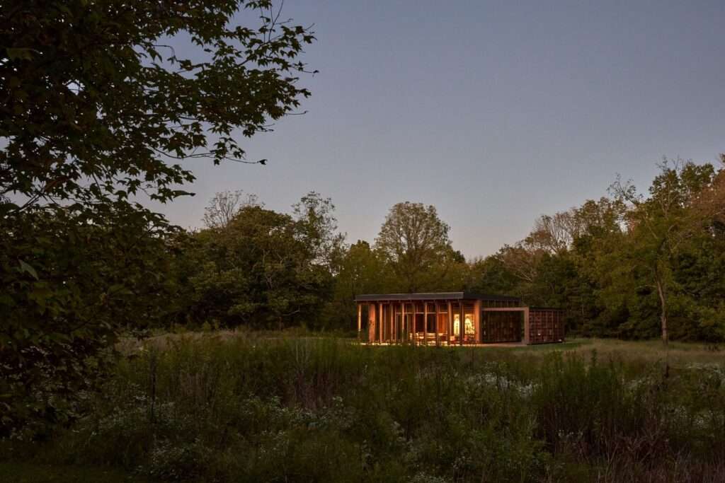 The Rock Pavilion at dusk, showing a minimalist glass and dark wood structure illuminated from within, situated on the edge of a dense forest and a wild grassy meadow under a twilight sky.
