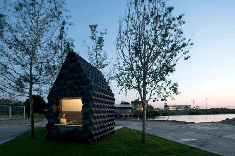 The 3D-printed Urban Cabin illuminated from the inside at dusk with a person visible through the large window.