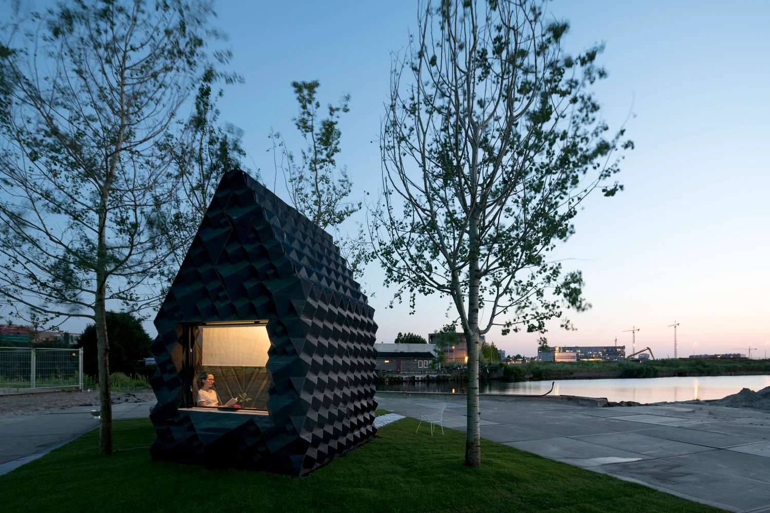 The 3D-printed Urban Cabin illuminated from the inside at dusk with a person visible through the large window.