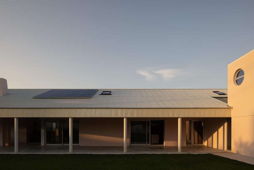 Architectural twilight shot of a continuous southern-facing veranda supported by concrete columns, showing warm interior light through glass doors, a corrugated metal roof with solar panels, and a circular window on the intersecting block.