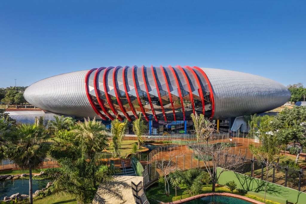 Aerial view of the Pantanal Biopark showing its iconic oval-shaped structure, red metallic frame, and zinc-clad roof in Campo Grande.