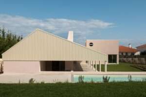 Modern L-shaped residential house with a prominent corrugated pitched roof, light pink stucco walls, concrete columns supporting a continuous veranda, and an outdoor swimming pool under a blue sky.