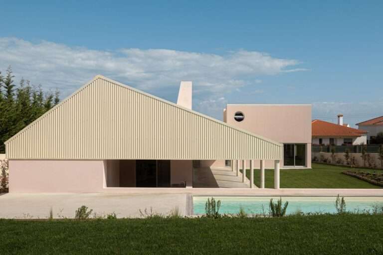 Modern L-shaped residential house with a prominent corrugated pitched roof, light pink stucco walls, concrete columns supporting a continuous veranda, and an outdoor swimming pool under a blue sky.