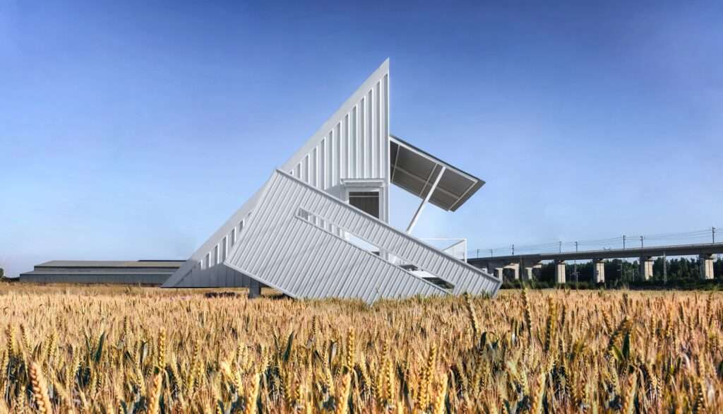 A geometric white metal rest house with a sharp triangular profile standing in a golden wheat field under a blue sky.