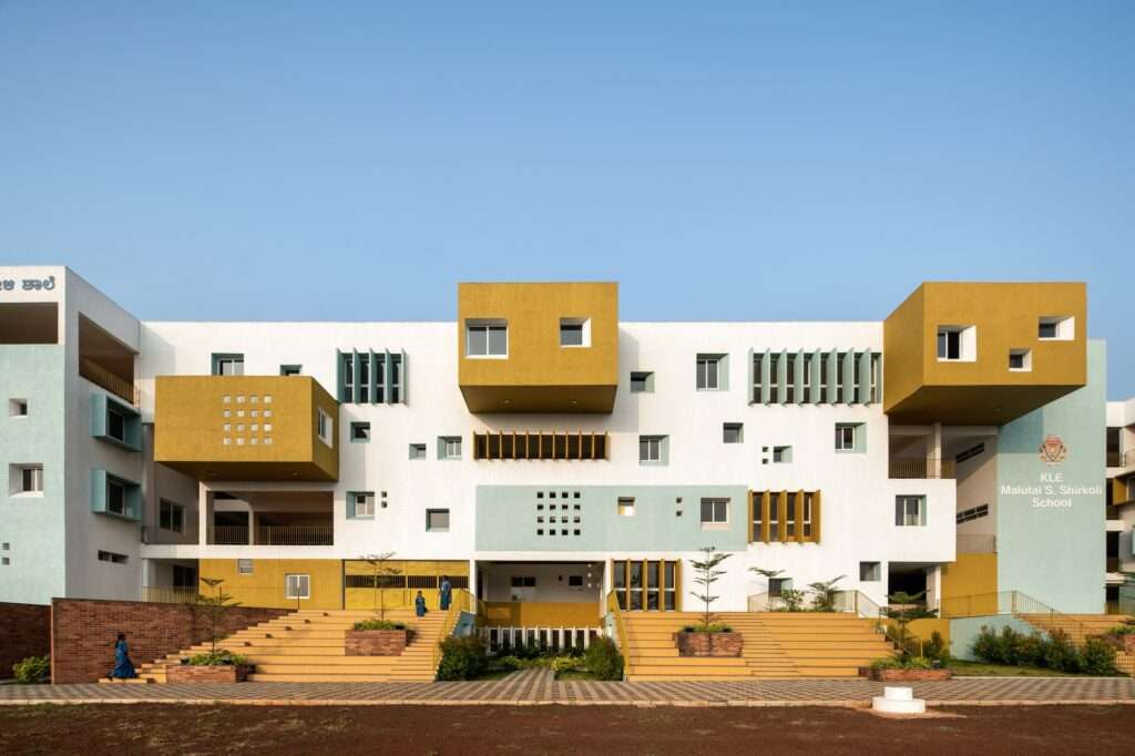 Modern school front facade with cantilevered mustard yellow blocks, asymmetric windows, and monumental wide concrete steps integrated with planters under a clear blue sky.