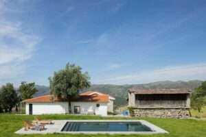 Ground-level view of Abadim House showing a minimalist rectangular swimming pool, wooden lounge chairs, a white plastered facade with a clay tile roof, and an adjacent historic stone outbuilding under a bright blue sky.