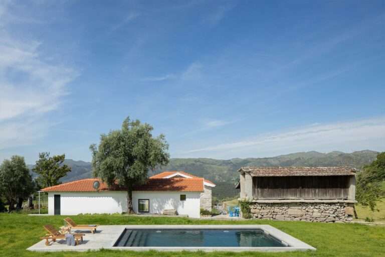 Ground-level view of Abadim House showing a minimalist rectangular swimming pool, wooden lounge chairs, a white plastered facade with a clay tile roof, and an adjacent historic stone outbuilding under a bright blue sky.