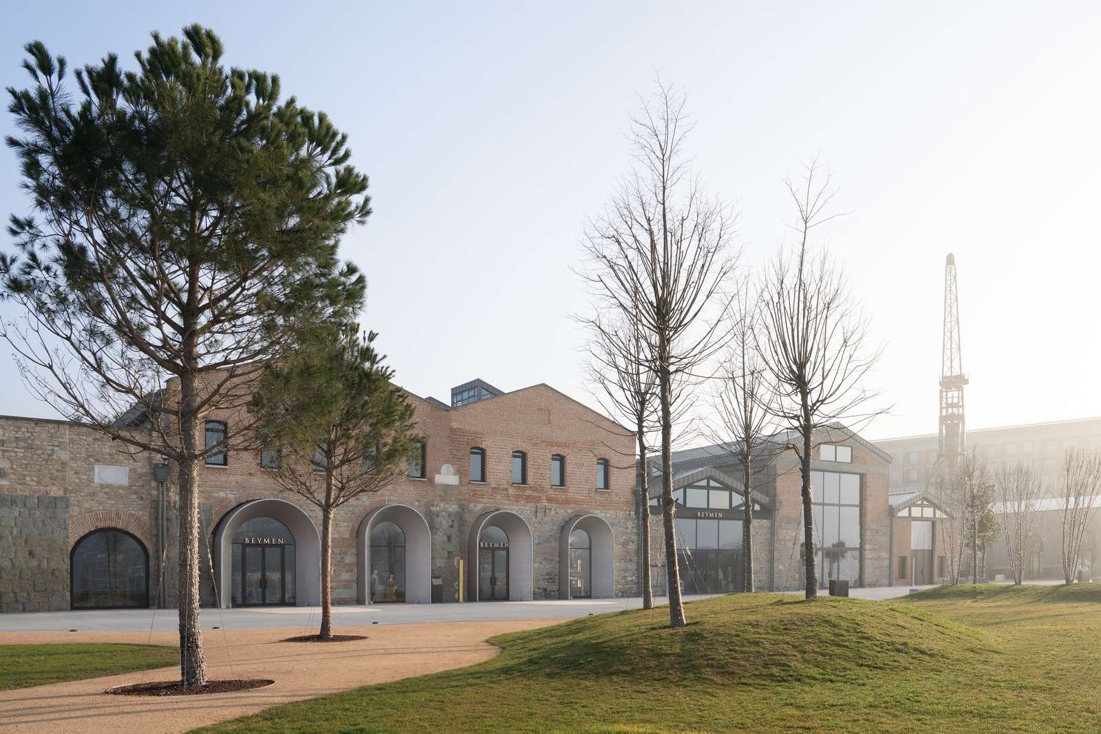 Exterior view of the brick facade of Beymen Tersane with historical arched entryways and a landscaped green plaza under a clear sky.