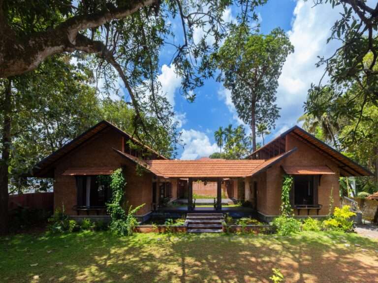 Front elevation of Shala Twam by Bhoomija Creations featuring red brick walls, traditional clay tiled roofs, and a central lily pond entry under lush green trees.