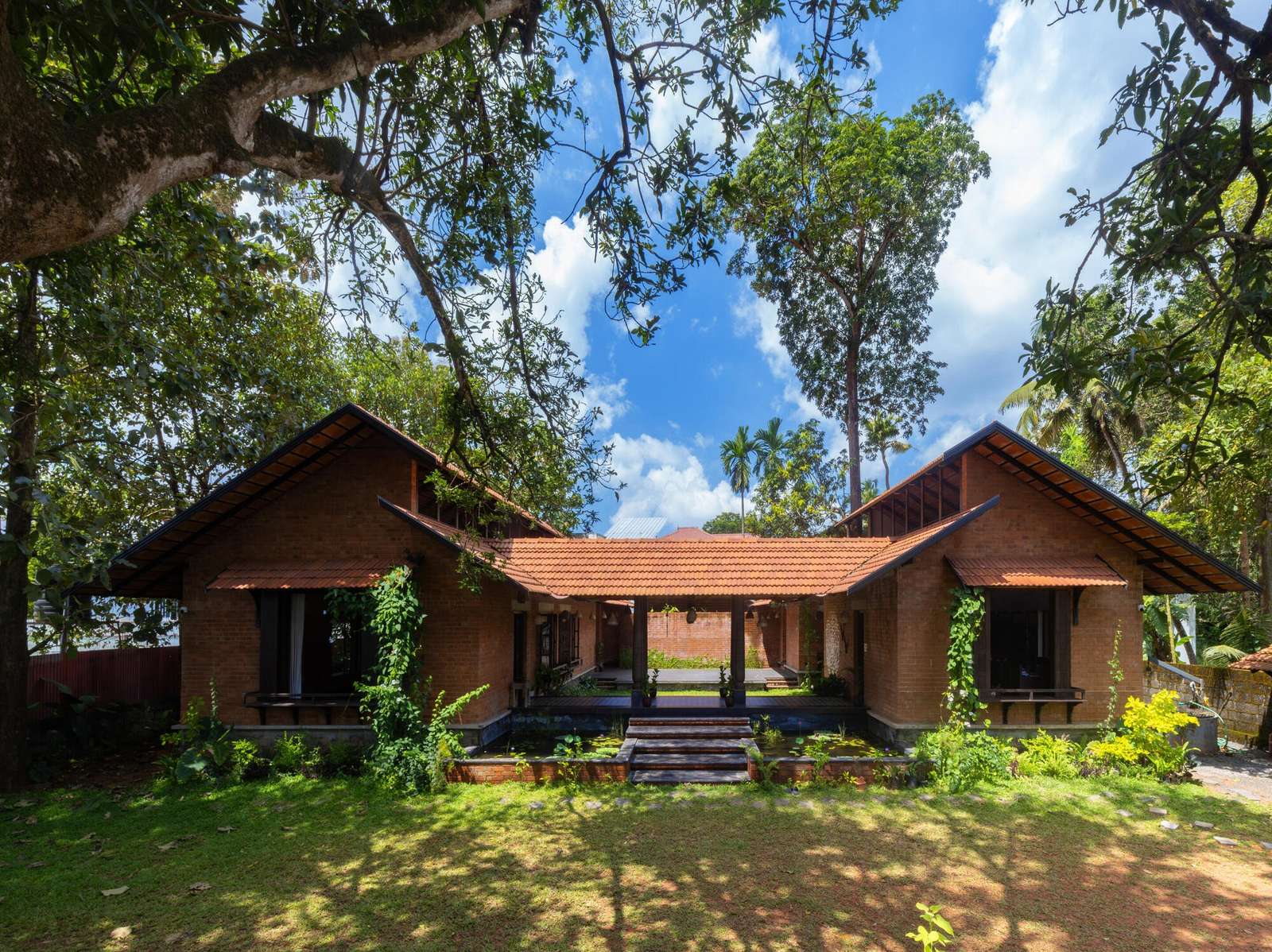 Front elevation of Shala Twam by Bhoomija Creations featuring red brick walls, traditional clay tiled roofs, and a central lily pond entry under lush green trees.