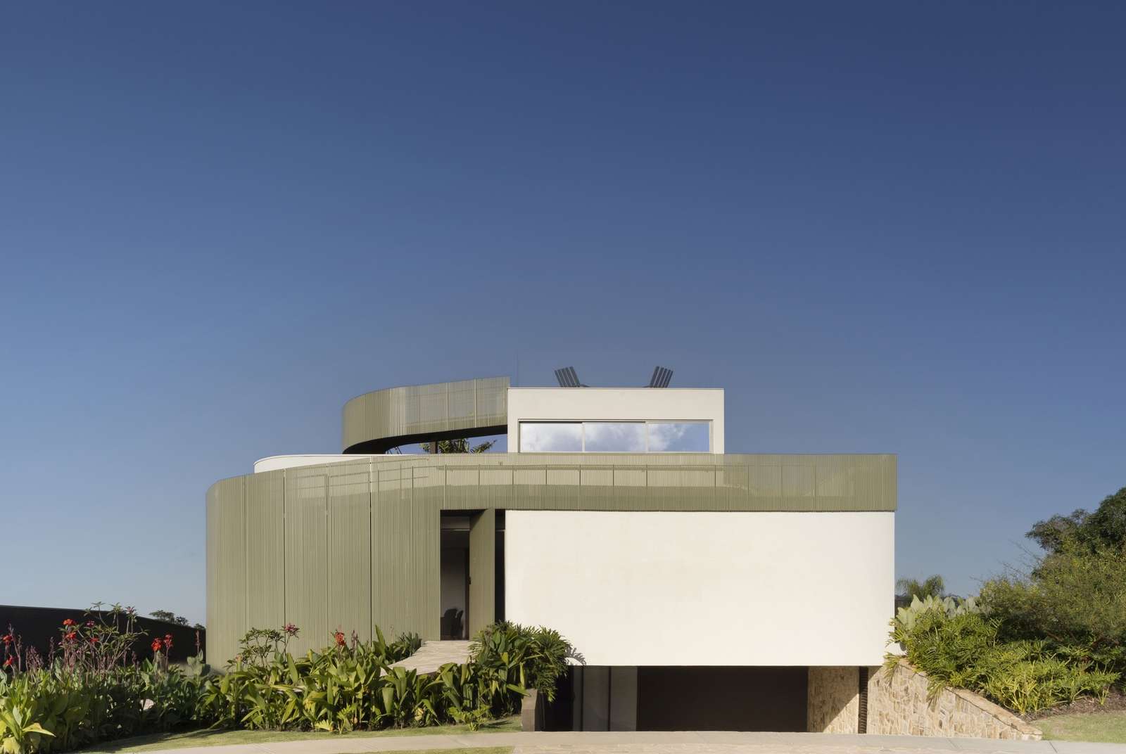 Front facade of Casa Circular showing the integrated ramp system, semi-transparent green metal louvers, lower-level garage entrance, and surrounding lush green landscaping under a clear blue sky.