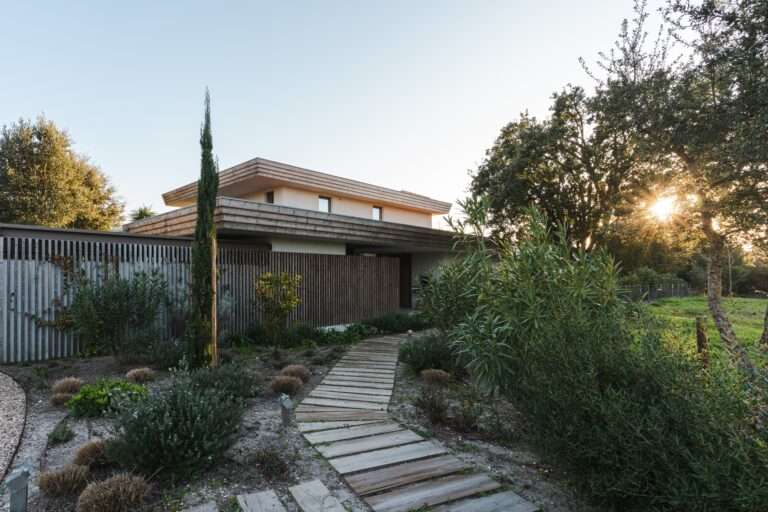 Wooden pathway leading to the modern residential entrance of Casa da Encosta with local vegetation and sunset light.