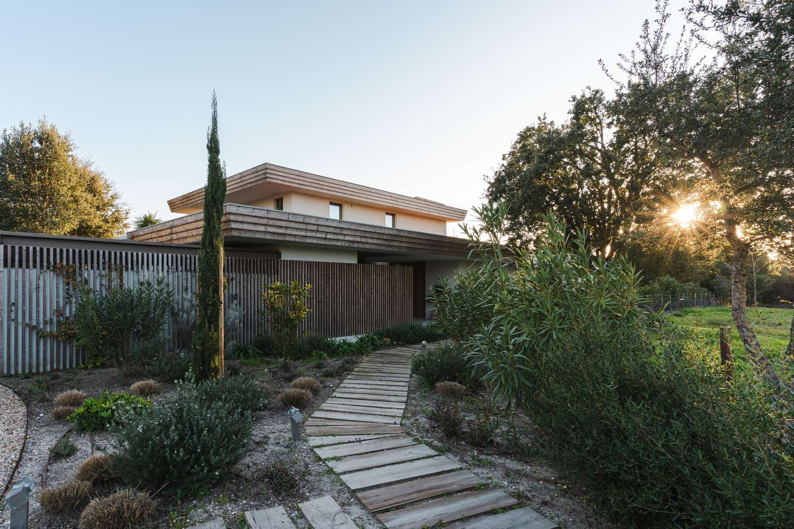 Wooden pathway leading to the modern residential entrance of Casa da Encosta with local vegetation and sunset light.