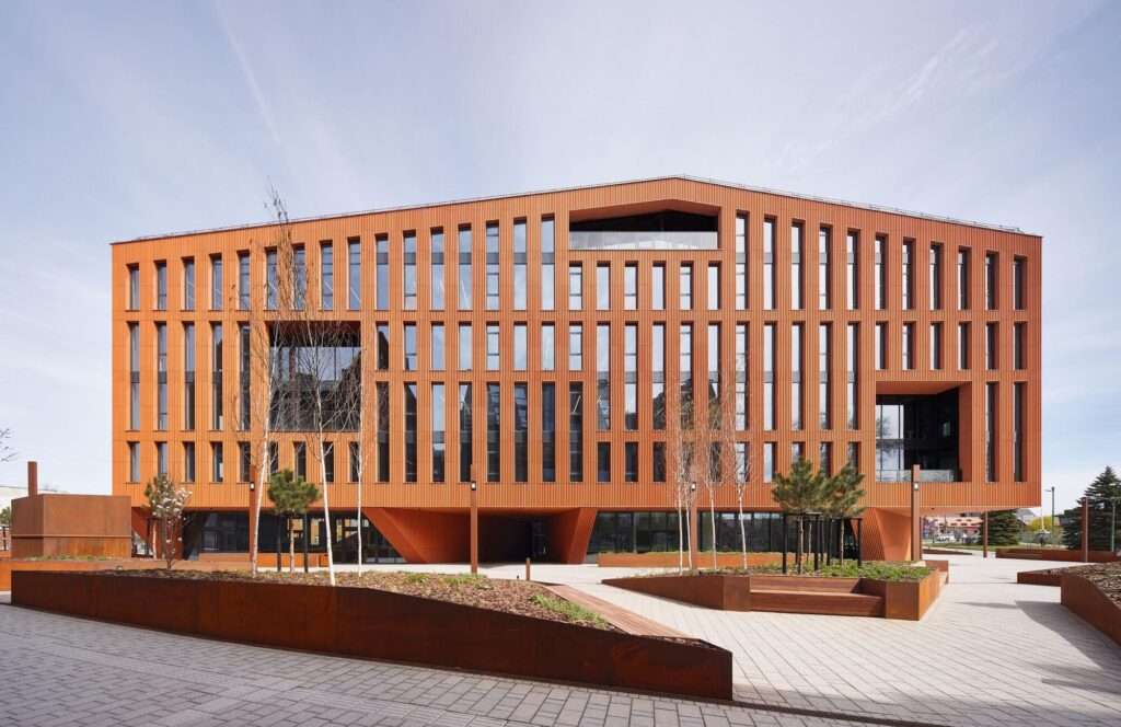 Front view of the HANZA building facade in Klaipeda featuring textured terracotta-colored vertical louvers, a sloped roofline, large rectangular recessed openings, and an open ground floor public plaza.