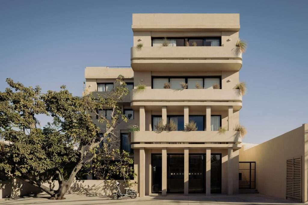 Front facade of Estero Residences showing a multi-story modern building with microcement finish, ground floor glass doors for Gamba Cafe, and integrated planters on cantilevered balconies.