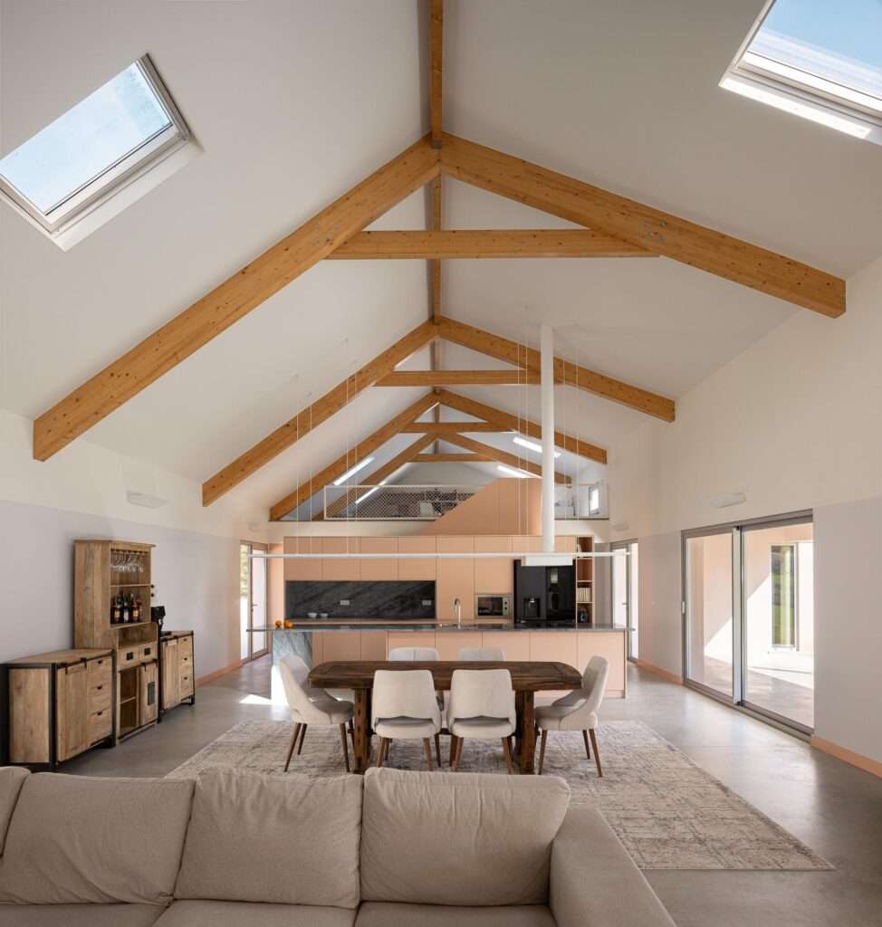 Spacious open-concept interior featuring a high gabled ceiling with exposed wooden trusses, skylights, a dining table with chairs, an integrated kitchen island, and large sliding glass doors opening to a courtyard.