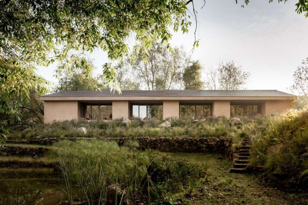 Monochromatic plastered pavilions of Rancho CP next to a natural lake with white horses running by the shore.