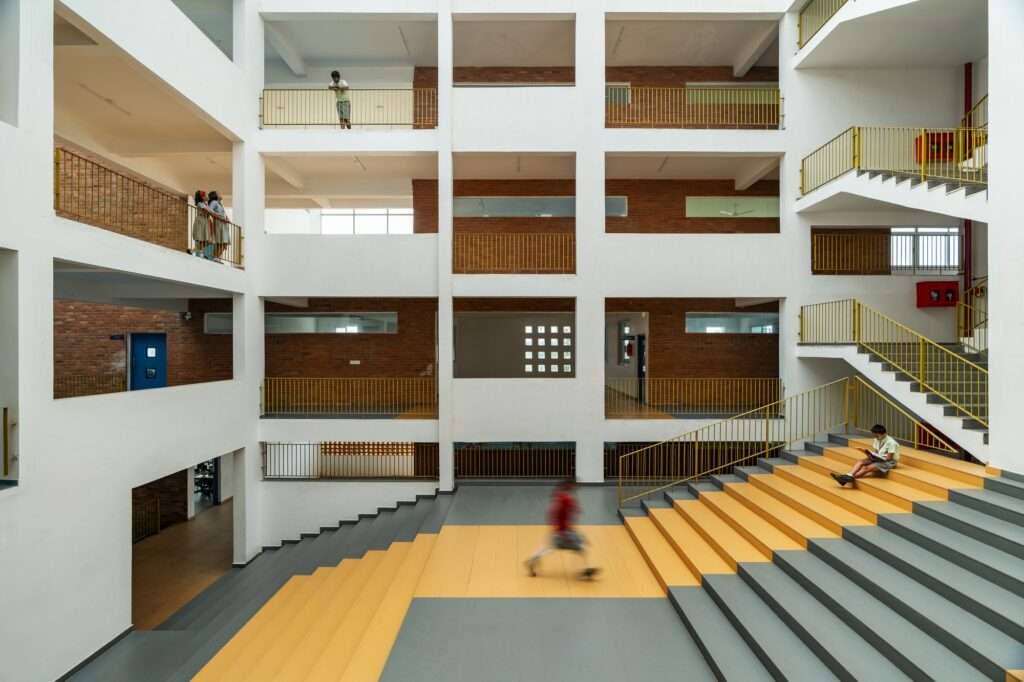Multi-story open-air atrium inside a contemporary school with wide yellow and grey steps where a student sits reading, overlooked by internal corridors with brick walls and yellow railings.