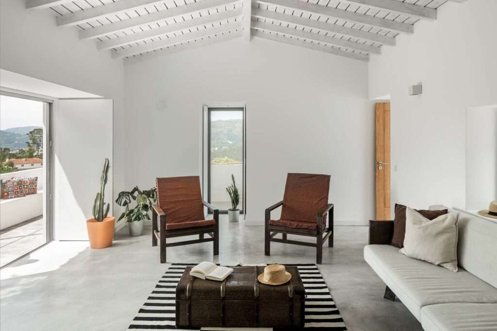 Bright contemporary living room interior featuring white painted wooden rafters, polished concrete flooring, rustic wooden armchairs, and minimalist decor opening to an outdoor terrace.