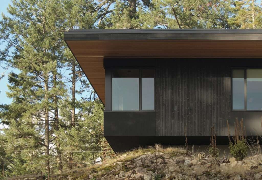 Side view close-up of Hilltop House cantilevered dark timber mass over a rocky stone terrain with evergreen pine trees.
