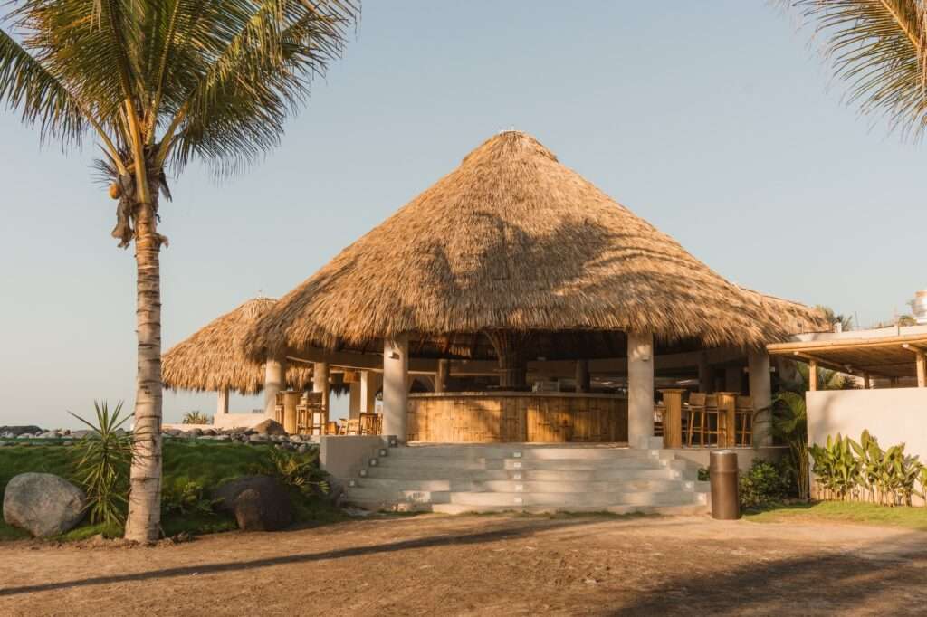 An outdoor beach bar with a large conical thatched palapa roof, supported by concrete columns and wrapped in a bamboo counter, set against a clear coastal sky with a palm tree.