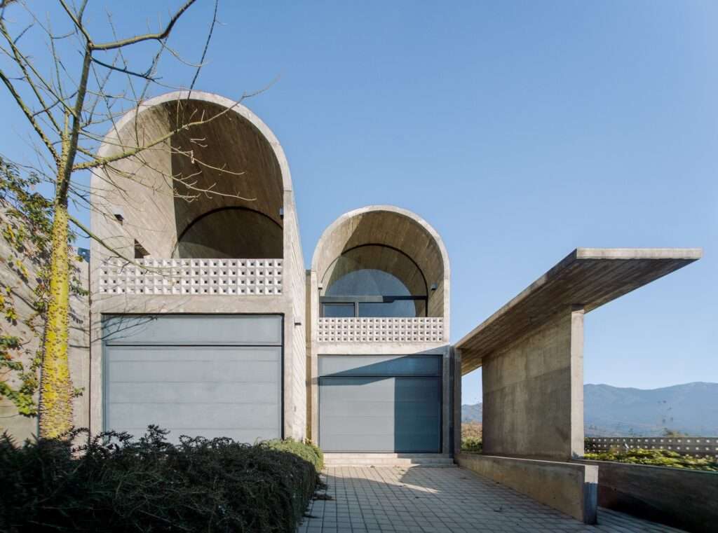Low-angle view of a textured raw concrete wall and cantilevered roof canopy at Casa Cielo above wild grasses and green landscaping.